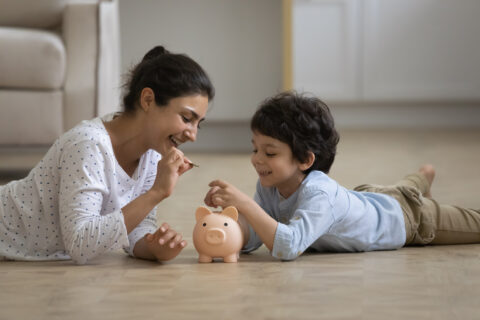 indian mother teaching little kid son saving money, putting coins in piggybank, lying on heated floor in Texas